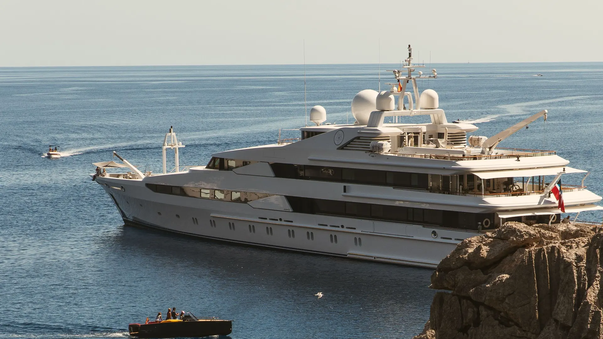 A white superyacht at anchor beside a rocky cliff in a blue bay